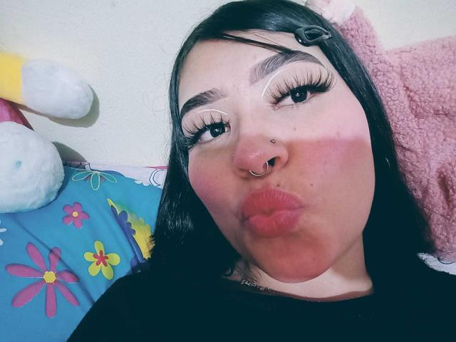 JulietaVelvet with black hair, nose ring, and long eyelashes, pouting her full, pink lips. Her eyes are half-open with thick, long lashes. She's wearing a black top.