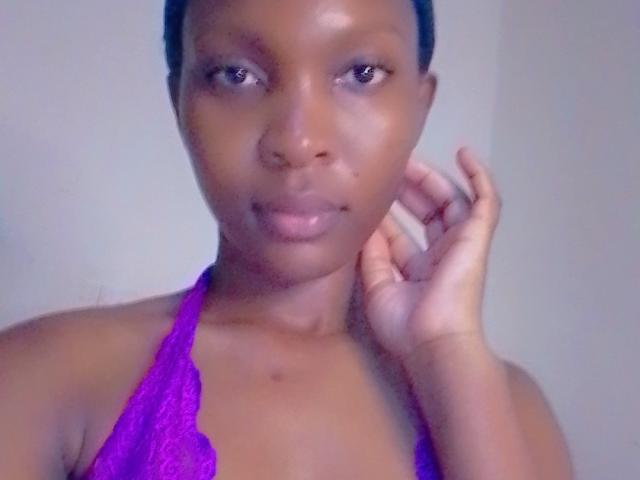 DinaNina has dark brown skin, shiny face, small mole below her lip, wearing a purple lace halter top, right hand touching her neck, short black hair, looking at camera.