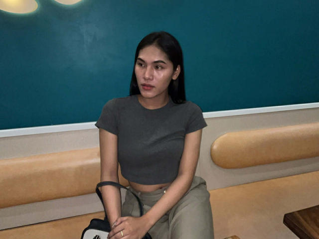 SofiaCumness with medium tits, dark brown hair, and light brown skin, wearing a gray crop top and beige pants, sitting on a tan bench, looking slightly annoyed.