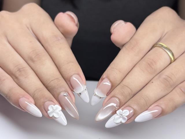 SofiaCumness's hands with white and pink nail art, featuring flowers and silver accents, wearing a gold ring, showing off long, sharp, almond-shaped nails.