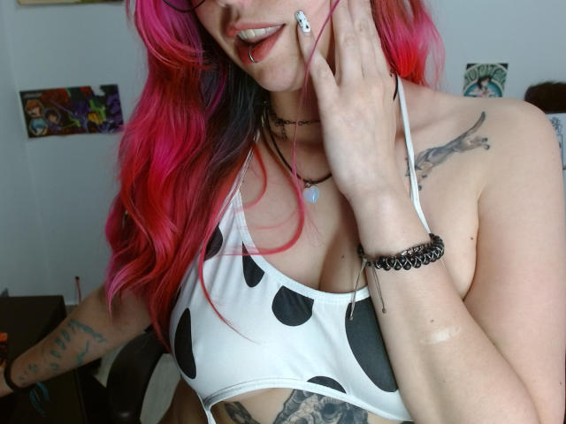 NatashaLau with pink and black hair, wearing a white polka dot bikini top, showing her tits and tattoos, holding her face, black choker, and bracelet, lip ring.