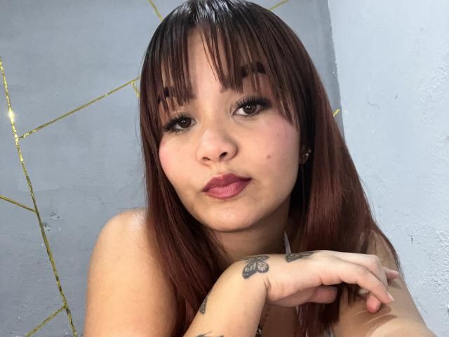 EstefyChaud has brown hair, bangs, and tattoos on her right hand, with her pouty lips slightly parted. She's looking at the camera with a subtle, sexy smile.