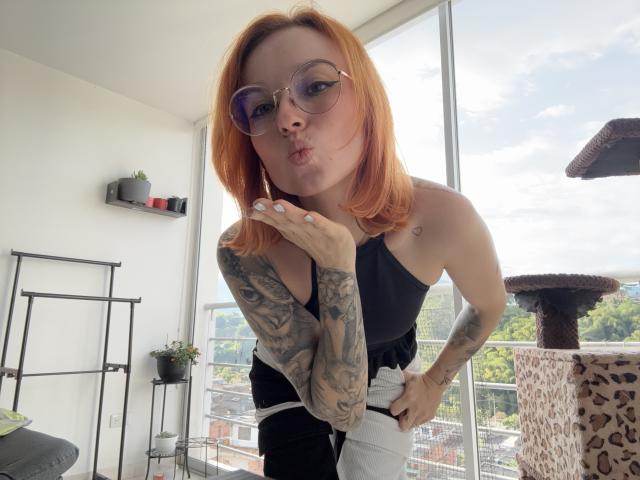 SarahRousen with orange hair, tattoos on arms, pouting lips, wearing glasses, black and white outfit, bending forward, hands near waist, standing in sunlit room with cat tree.