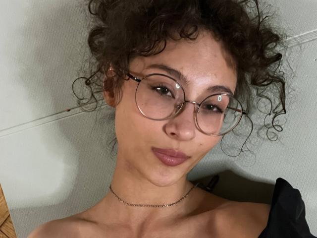 AlyceErway with curly brown hair, wearing round glasses, a thin necklace, and a black strap, has puffy lips and a slight smirk, showing her tits.