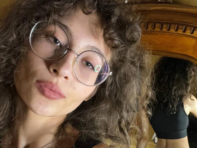 AlyceErway with curly brown hair, wearing round glasses, pink lipstick, and a black top, showing a mirror reflection of her bare back. Boobs not visible. Reflective glasses.
