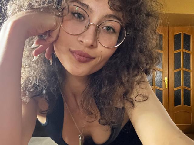 AlyceErway with curly brown hair, wearing glasses, a black tank top, and a necklace, has puffy pink lips and is touching her cheek, showing her tits.
