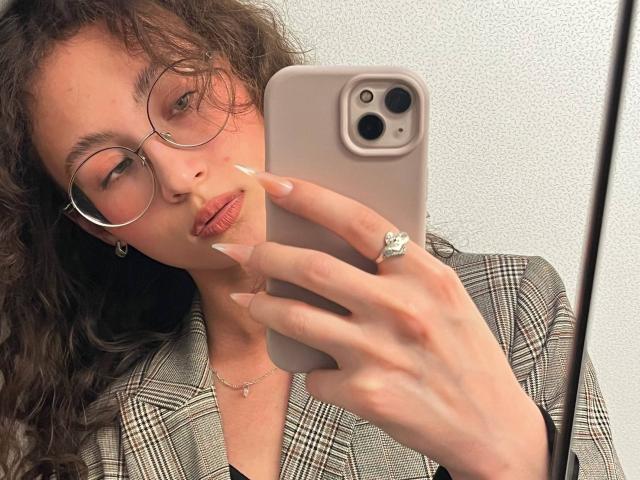 AlyceErway with curly brown hair, glasses, and long nails, holding a phone with a diamond ring, wearing a plaid shirt, taking a mirror selfie.