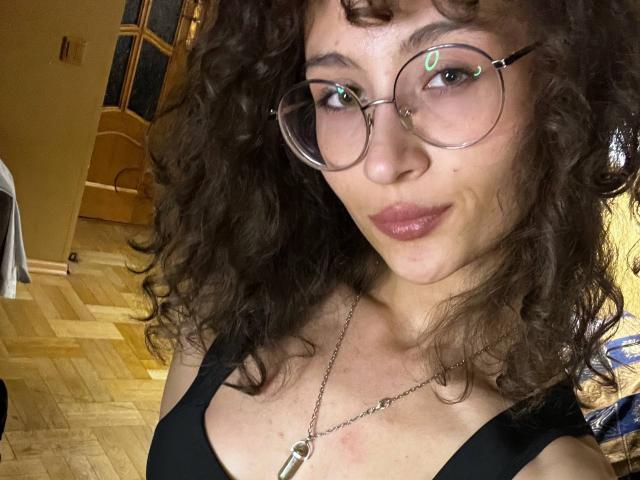 AlyceErway with curly brown hair, glasses, and a nose ring, wearing a black top, showing her boobs, and a necklace, smiling slightly in a selfie.