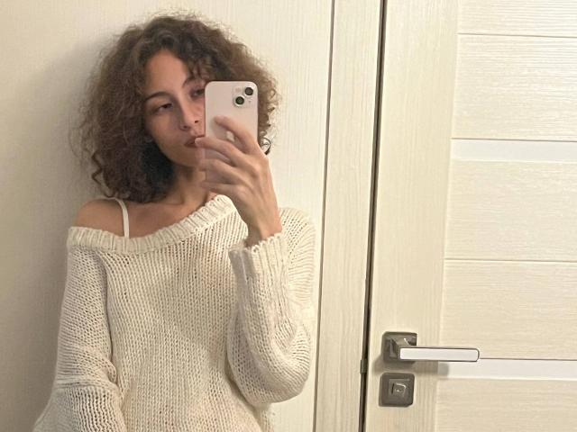 AlyceErway with curly brown hair, sucking her finger, wearing a white off-shoulder knit sweater, taking a mirror selfie with a pink iPhone.