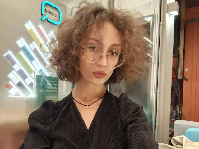 AlyceErway with curly brown hair, wearing round glasses and a black pinstriped shirt, has a necklace and a serious expression, standing in a brightly lit room.