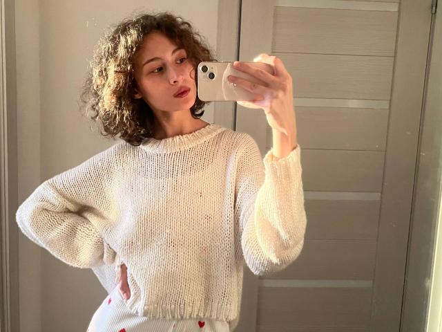 AlyceErway with curly brown hair, wearing a white knit sweater, taking a mirror selfie with her hand on her hip, showing off her ass in white pants.