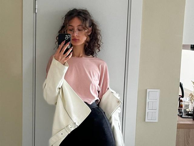 AlyceErway with curly brown hair, pink top, white jacket, and black high-waisted pants, holding a phone, standing in front of a door, showing her ass.
