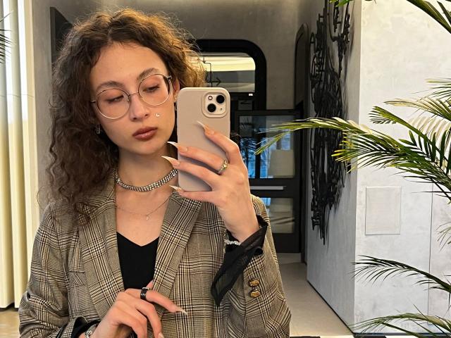 AlyceErway with curly brown hair, glasses, and nose ring, wearing a plaid blazer, black top, and chain necklace, taking a mirror selfie with a gold phone.