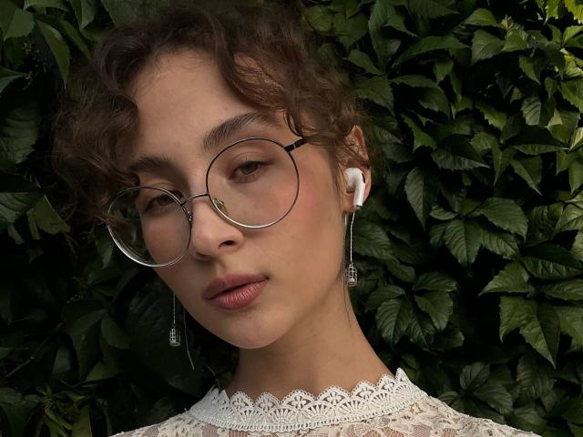 AlyceErway with curly brown hair, round glasses, white lace collar, wearing earbuds, has light brown skin, full pink lips, and a serious expression. Green leafy background.