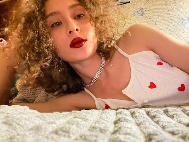 AlyceErway with curly blonde hair, red lipstick, white ribbed tank top with red hearts, diamond necklace, lying on a textured gray blanket, looking at the camera.