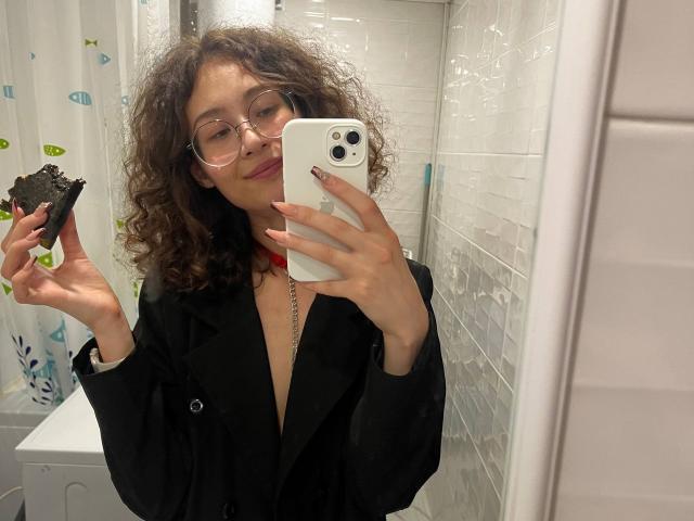 AlyceErway with curly brown hair, glasses, and a black jacket, holding a dark rock in a white-tiled bathroom, taking a selfie with an iPhone.