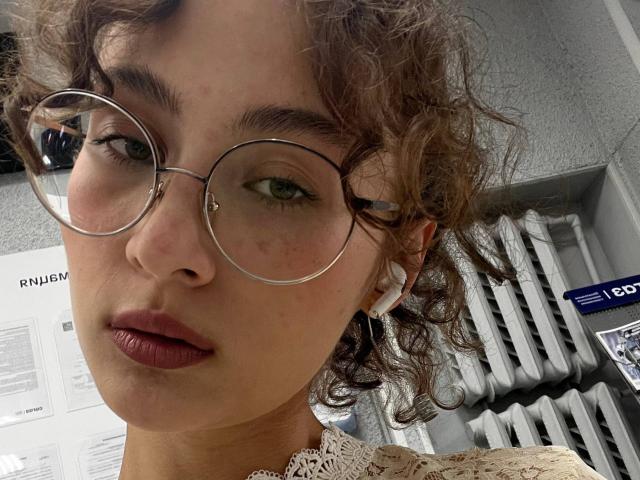 AlyceErway with curly brown hair, wearing round glasses, white earbuds, and a lace top, has puffy pinkish lips and light freckles, in a soft-focus selfie.