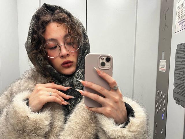 AlyceErway with curly brown hair, round glasses, and a fur coat, takes a selfie in an elevator. She's wearing a gray scarf and a ring on her right hand.
