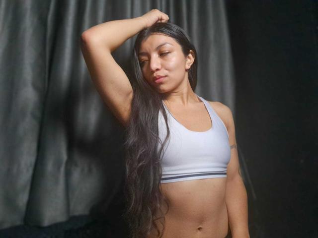 IbbyeMoreau with long black hair, eyes closed, arm raised, wearing a white sports bra, showing her small tits and toned abs, standing in front of dark curtains.