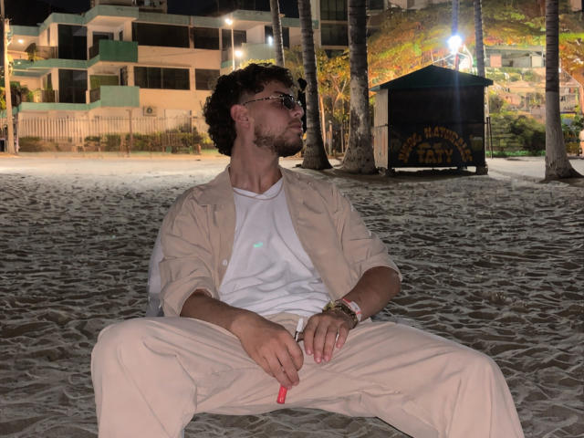 Sevven, with curly dark hair and beard, wearing sunglasses, beige shirt, white tee, and beige pants, sits on sandy beach at night, holding red cigarette, relaxed, right hand on thigh.