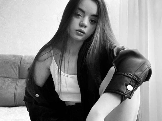 AnasteishaXXX-hot with long hair, wearing a white tank top and black jacket, sits with one leg up, showing a leather arm sleeve, in a black-and-white photo.