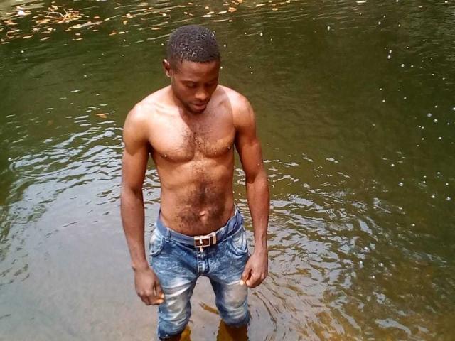 Un hombre negro, sin camisa, con el pecho peludo y la piel morena, está de pie en aguas poco profundas, vistiendo unos vaqueros azules, ligeramente mojados, con un cinturón marrón, y mirando hacia abajo..