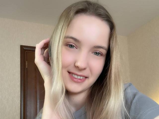 NicoleSpritzer with blonde hair, green eyes, and pink lips, smiling softly, holding a phone to her ear, wearing a gray shirt, with a cute, natural face.
