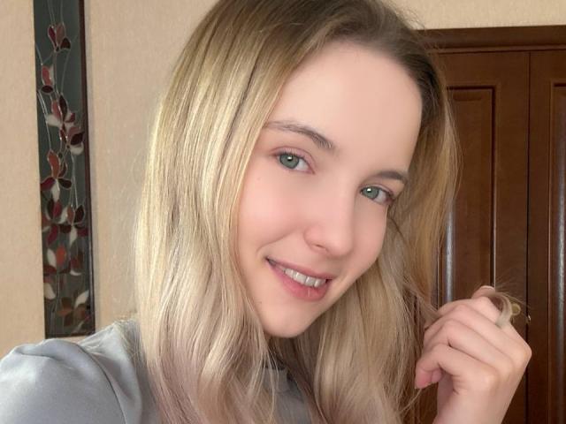 NicoleSpritzer has blonde hair, green eyes, and a cute smile with her hand in her hair. She's wearing a grey shirt, and her tits are small and natural.