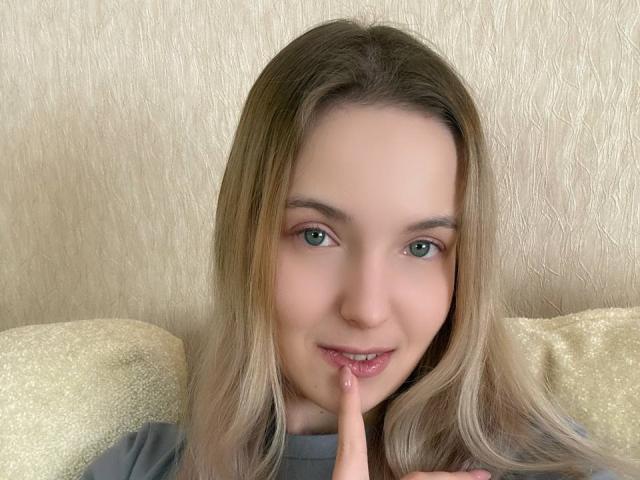 NicoleSpritzer has light brown hair, green eyes, and a cute smile, with her finger touching her pink lips. She's wearing a gray shirt and lying on a beige pillow.