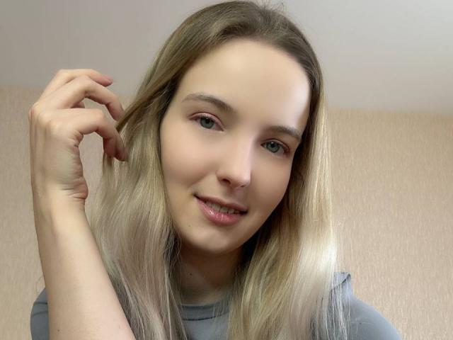 NicoleSpritzer has light blonde hair, blue eyes, and a slight smile with her hand touching her hair. She's wearing a gray shirt and has a cute face.