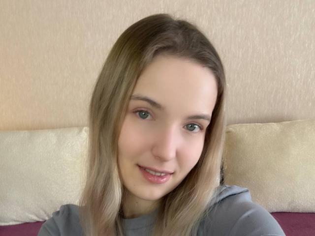 NicoleSpritzer has light blonde hair, green eyes, and a cute smile with pink lips. She's wearing a gray top, and her face is slightly turned to the right.