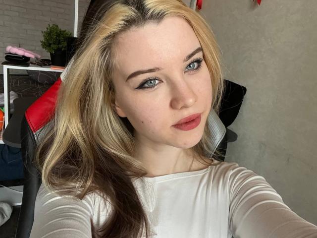 YolandoMysak with blonde and brown hair, blue eyes, and red lips, wearing a white top, sitting in a chair, looking at the camera with a slight pout.