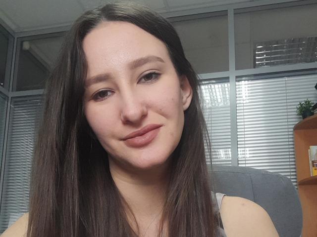 Omella's got a cute face with long brown hair, subtle makeup, and a small smile. She's showing off her tits in a grey tank top, sitting in an office.