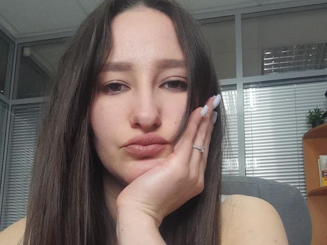 Het gezicht van Omella, met lang bruin haar, lichtjes opgetrokken lippen, een hand die haar wang aanraakt, een zilveren ring zichtbaar, witte nagels, die recht in de camera kijkt..