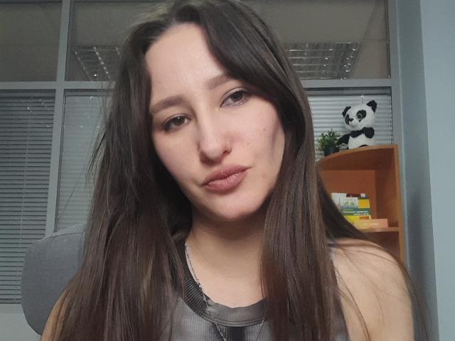 Omella has long brown hair, light skin, and is pouting with her lips slightly puckered. She's wearing a gray tank top, showing her small tits. Office background.
