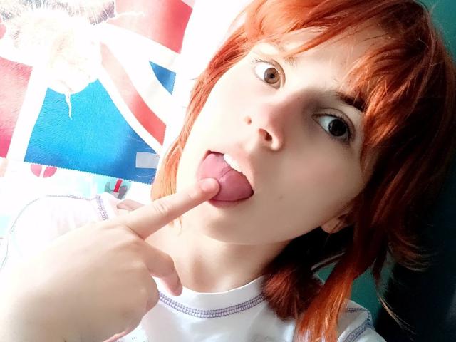 MireilleSpotwood with red hair, tongue out, sticking her finger in her mouth, brown eyes, white shirt, green wall in background. Her finger is touching her tongue.