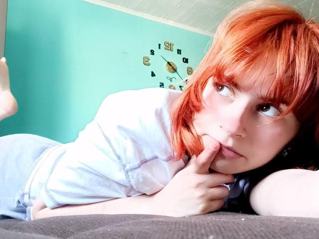 MireilleSpotwood with red hair, lying on a bed, wearing a white shirt, touching her chin with her finger, looking to the side, showing her ass in the foreground.
