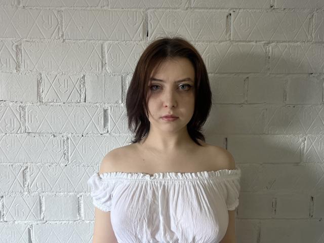 ValeryDuboise with shoulder-length brown hair, wearing a white off-shoulder top, showing her tits, standing against a textured white brick wall.