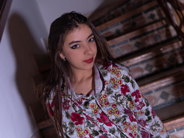 EvangelinaRosa with wet brown hair, floral dress, red lipstick, and blue eyeliner, standing on wooden stairs, looking up with a slight smile, showing her tits.