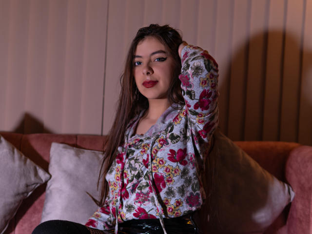 EvangelinaRosa with long brown hair, floral shirt, and black pants, hand in hair, red lipstick, sitting on pink couch with white pillows, looking at camera.