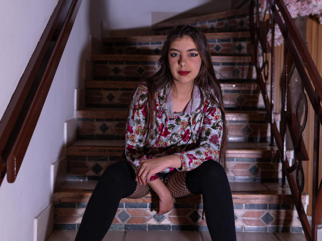 EvangelinaRosa with long brown hair, floral shirt, black leggings, and fishnet stockings, sitting on stairs, legs spread, looking at camera, red lipstick, wooden railings on both sides.