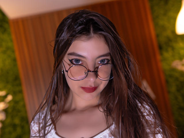 EvangelinaRosa with wet brown hair, round glasses, blue eyes, and red lipstick, wearing a white top, looking up at the camera, her small tits slightly visible.
