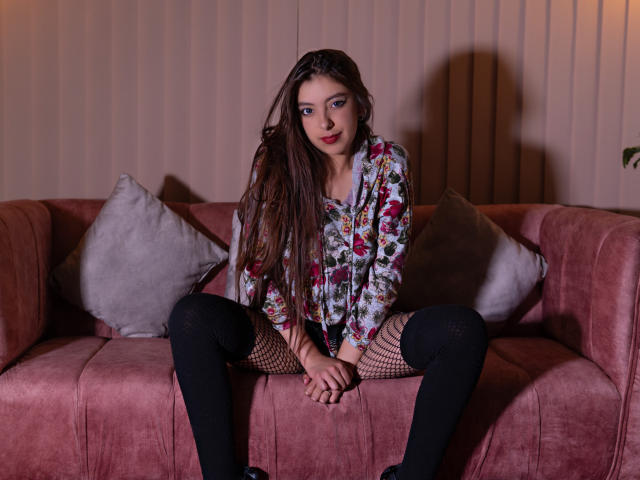 EvangelinaRosa sits on a pink couch, wearing a floral shirt, black fishnet stockings, and knee-high socks, with her legs spread, showing her pussy. Brown hair, blue eyes, red lips.