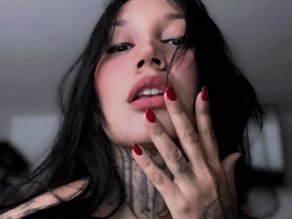 PaulinaV with black hair, red nails, tattoos on her neck, and slightly open mouth, touching her face. She's looking seductive with a slight blush on her cheeks.