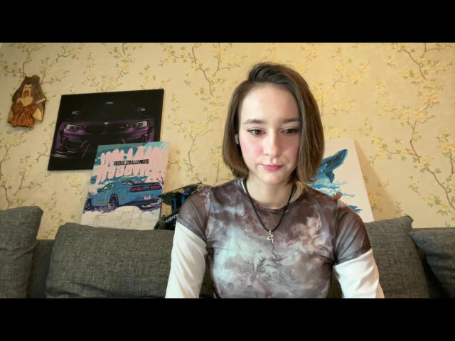 ZofiaWeigold with short brown hair, pink cheeks, wearing a tie-dye shirt, sitting on a grey couch, necklace, and wall art with cars in the background.