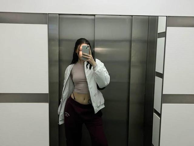 AlenaShimon with long black hair, wearing a beige crop top, white jacket, and maroon pants, taking a mirror selfie in an elevator, showing her small tits and flat ass.