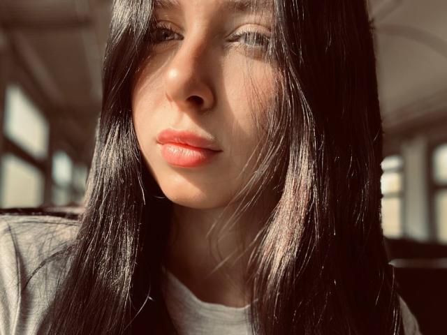 AlenaShimon has shiny black hair, sunlit skin, and red lips. She's wearing a gray shirt, and her eyes are slightly squinted from the sunlight.