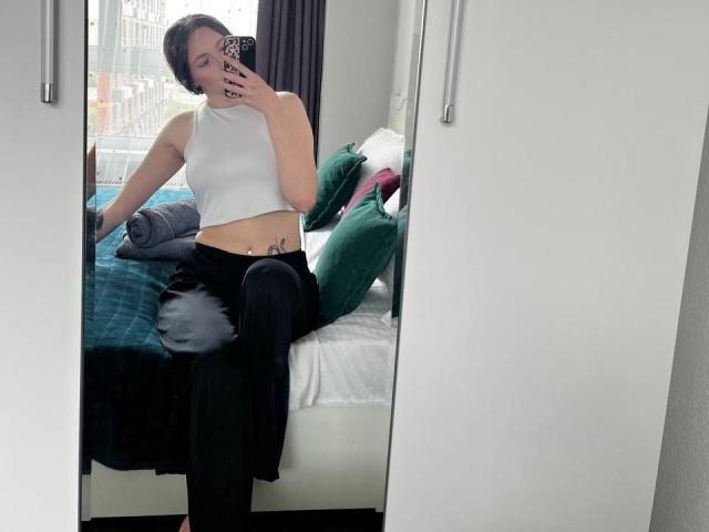 AlenaShimon with short dark hair, white crop top, black tie-dye pants, sitting on bed, taking mirror selfie, visible tattoo on right side, green pillows in background.