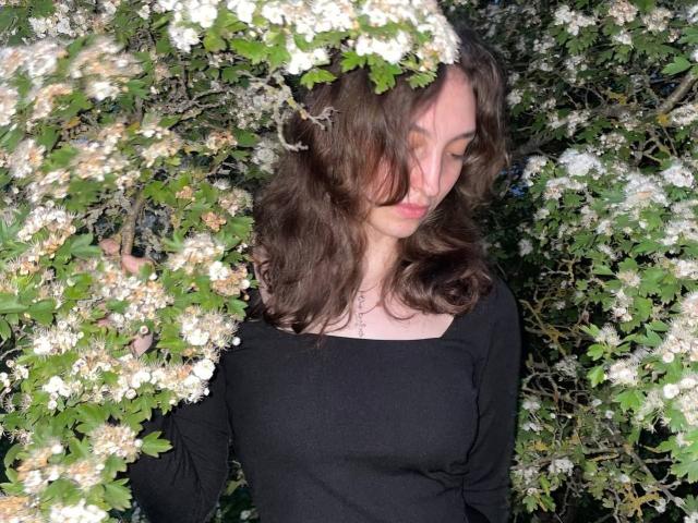 LeonieRahim with wavy brown hair, wearing a black top, surrounded by white flowers and green leaves, has her eyes closed, looking peaceful, with her hand in the foliage.