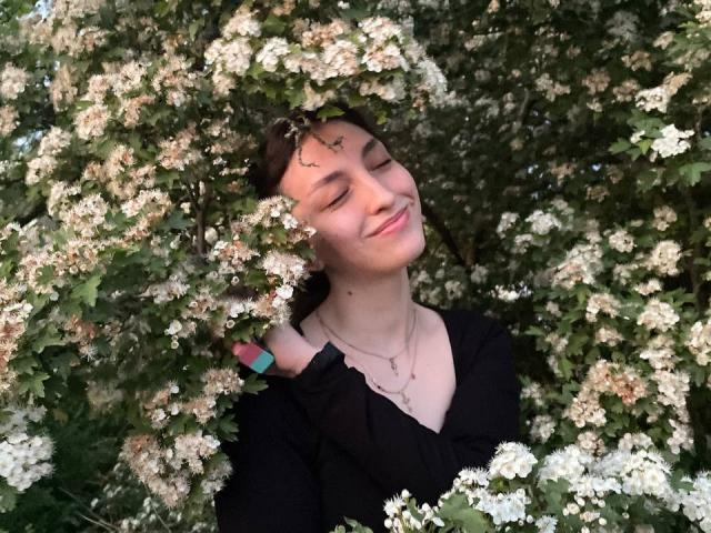 LeonieRahim with eyes closed, smiling, surrounded by white flowers, wearing black top, pink nail polish, blue nail polish, and necklace, looking happy, head in flower cluster.